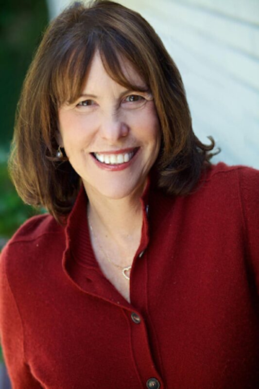 Smiling woman with shoulder-length brown hair, wearing a red jacket, standing in front of a light-colored wall.