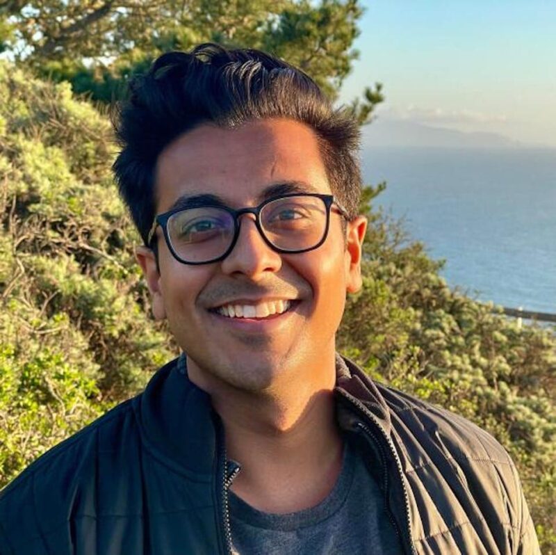 Smiling person wearing glasses and a dark jacket standing outdoors, with the ocean and greenery in the background.