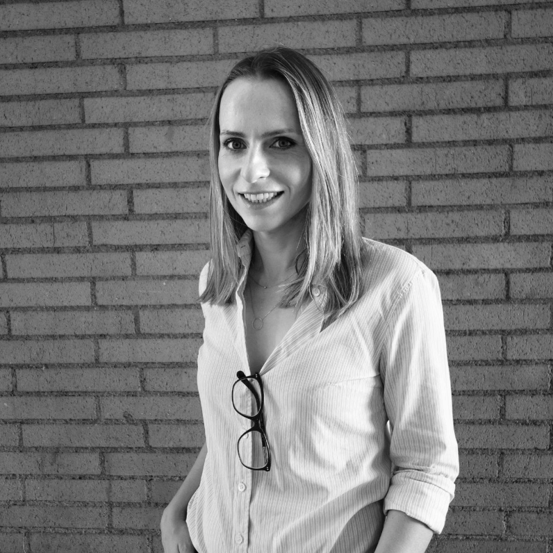 A person stands smiling against a brick wall, wearing a striped shirt with glasses hanging on the collar. Black and white photo.