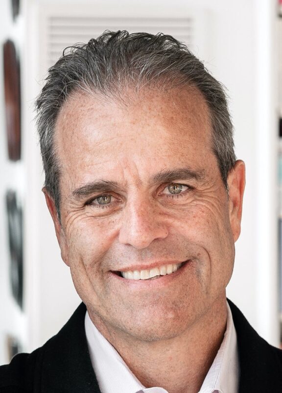 Smiling man with short gray hair wearing a black jacket and white shirt, standing indoors with a blurred background.