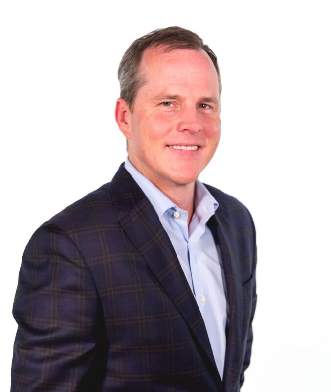 Man in a checkered blazer and blue shirt smiling against a white background.