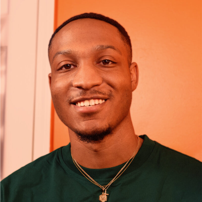 Smiling person wearing a green shirt and gold necklaces, standing against an orange background.