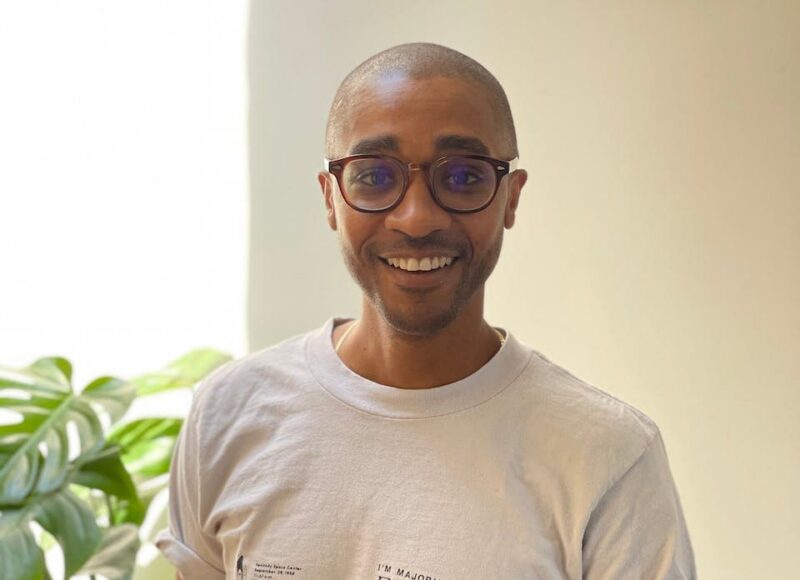A person with glasses smiles indoors, wearing a light-colored shirt. A green leafy plant is partially visible on the left.