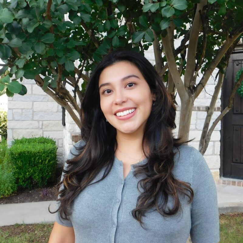 A smiling person with long dark hair stands outdoors in front of a tree and a stone building, wearing a gray shirt.