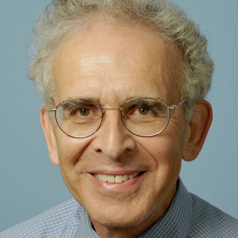 Elderly person with curly gray hair, wearing glasses and a blue shirt, smiles against a plain blue background.