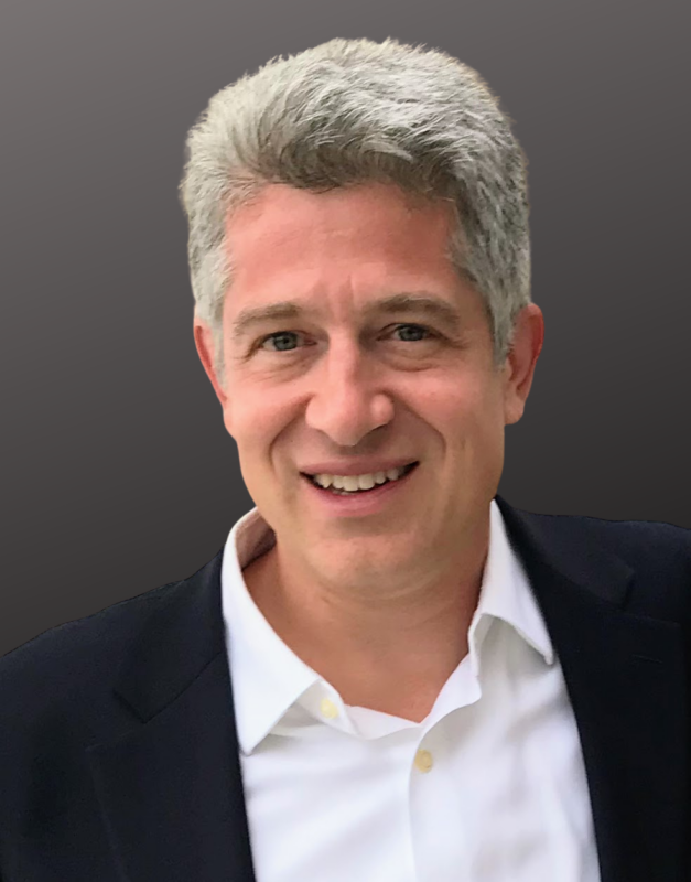 Smiling man with short gray hair wearing a white shirt and dark blazer against a dark background.