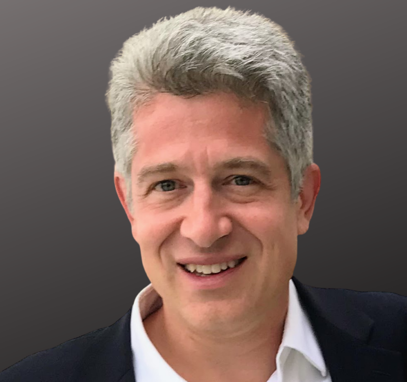 Smiling man with short gray hair wearing a white shirt and dark blazer against a dark background.