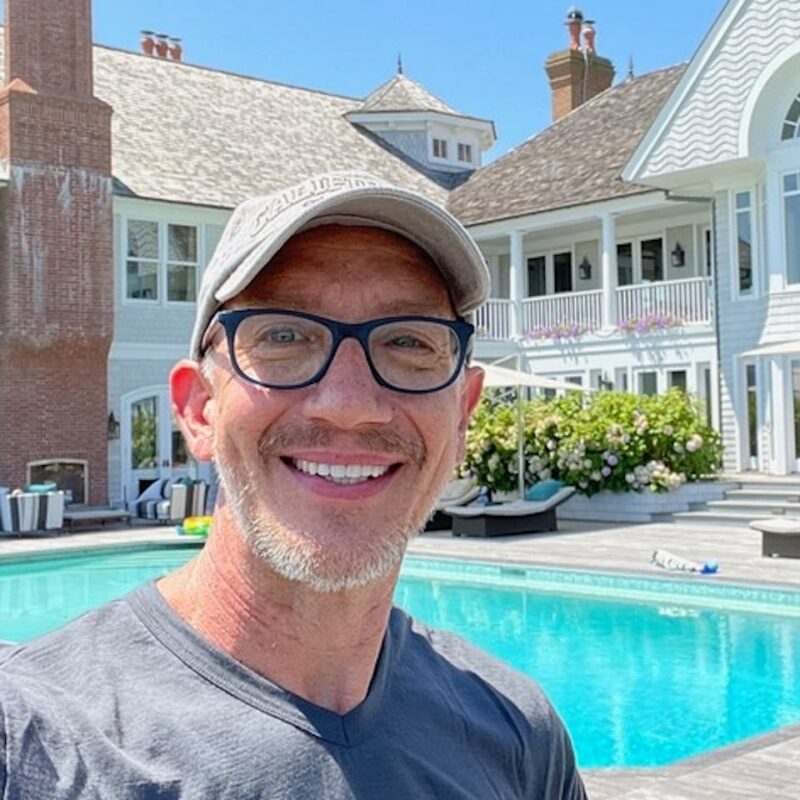 Man in glasses and cap smiling by a large pool, with a white multi-story house in the background under a blue sky.