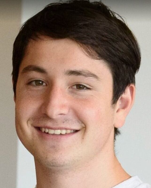 Smiling young man with short dark hair and light stubble, wearing a white T-shirt, stands against a blurred background.