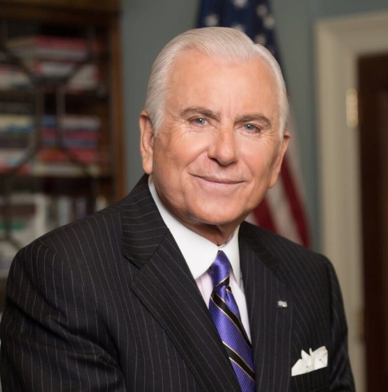 Smiling elderly man in a suit and tie stands in an office with bookshelves and a U.S. flag in the background.