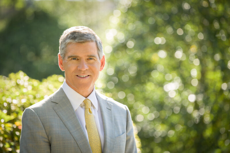 Man in a gray suit and yellow tie smiling outdoors, with a blurred green background of trees.