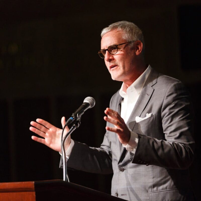 Man with gray hair and glasses in a suit speaks at a podium with a microphone, gesturing with both hands.