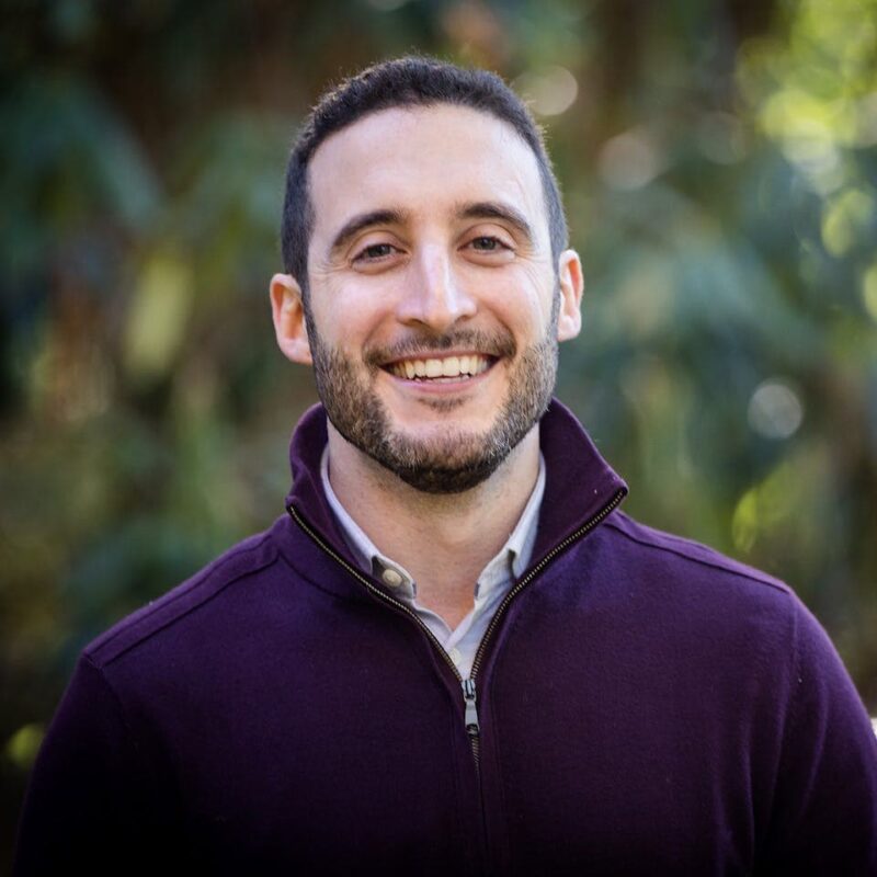 Man with short hair and beard smiling, wearing a maroon zip-up jacket outdoors with blurred greenery in the background.