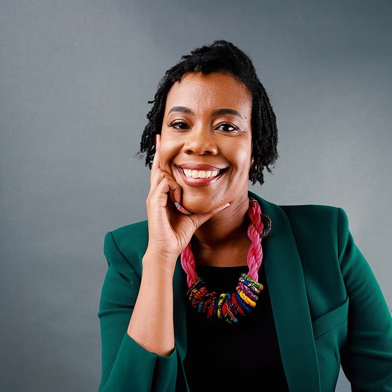 Smiling person with short hair, wearing a green blazer and colorful necklace, rests their chin on their hand against a gray background.