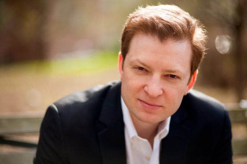 Man with short red hair in a black suit jacket and white shirt, sitting on a bench outdoors, looking at the camera with a neutral expression.