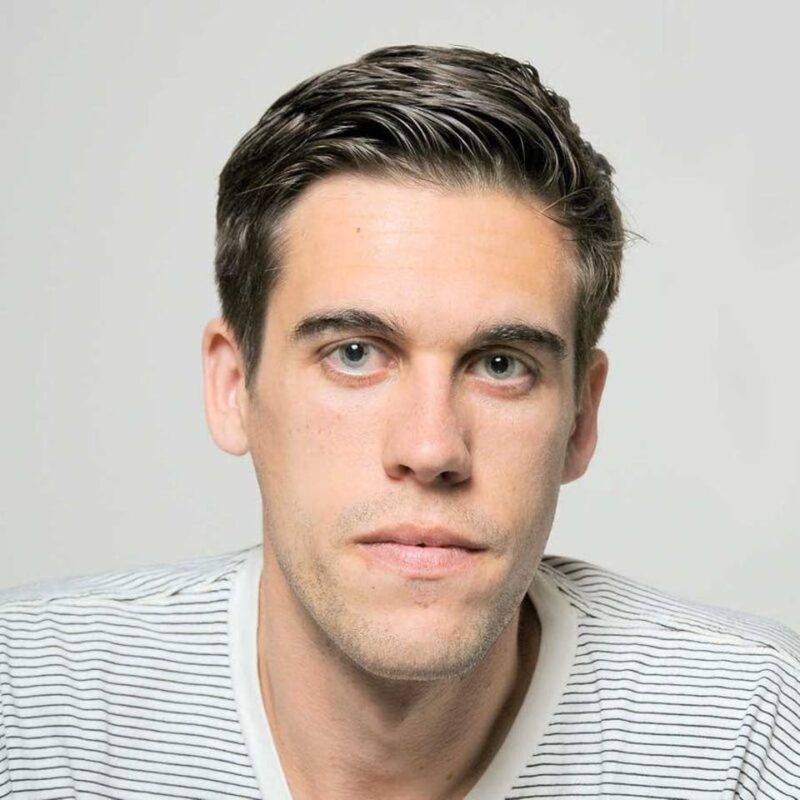 A person with short dark hair, wearing a white shirt with black stripes, looks directly at the camera against a neutral background.