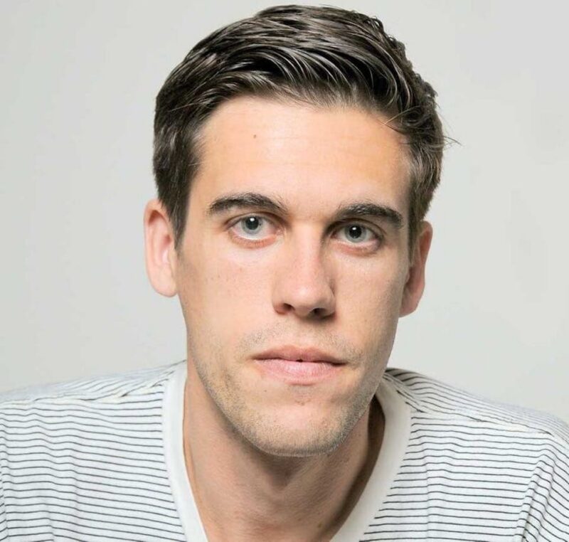 A person with short dark hair, wearing a white shirt with black stripes, looks directly at the camera against a neutral background.
