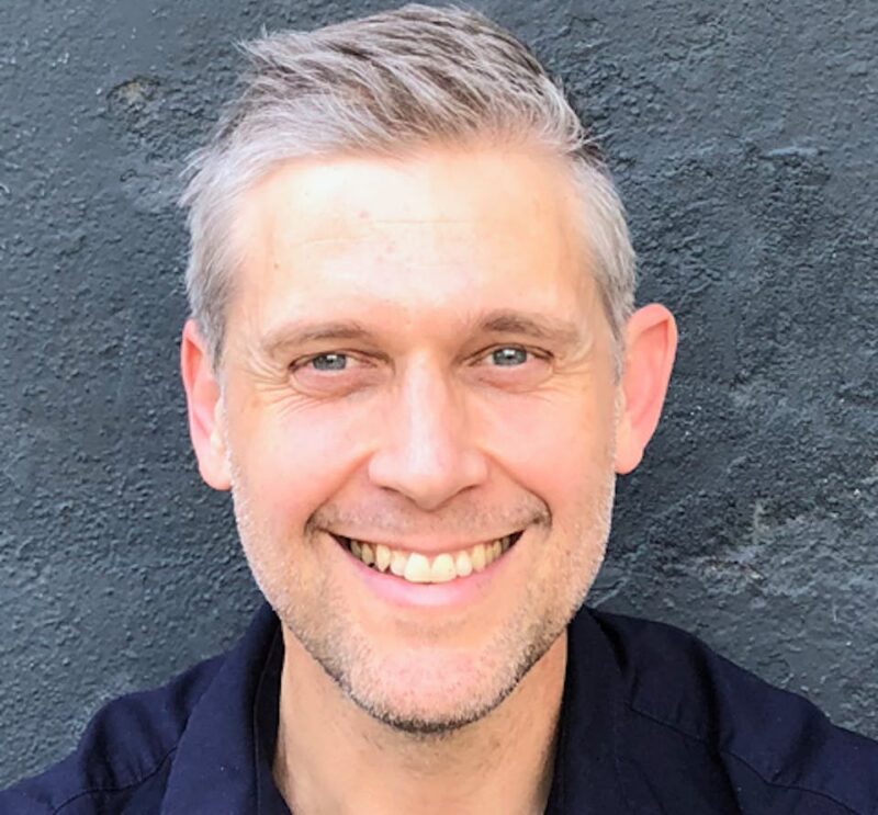 Smiling man with short gray hair and a black shirt against a dark textured wall.