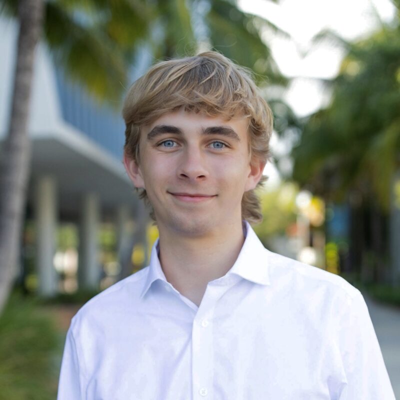 Person in a white shirt smiling outdoors, with palm trees and a building in the background.