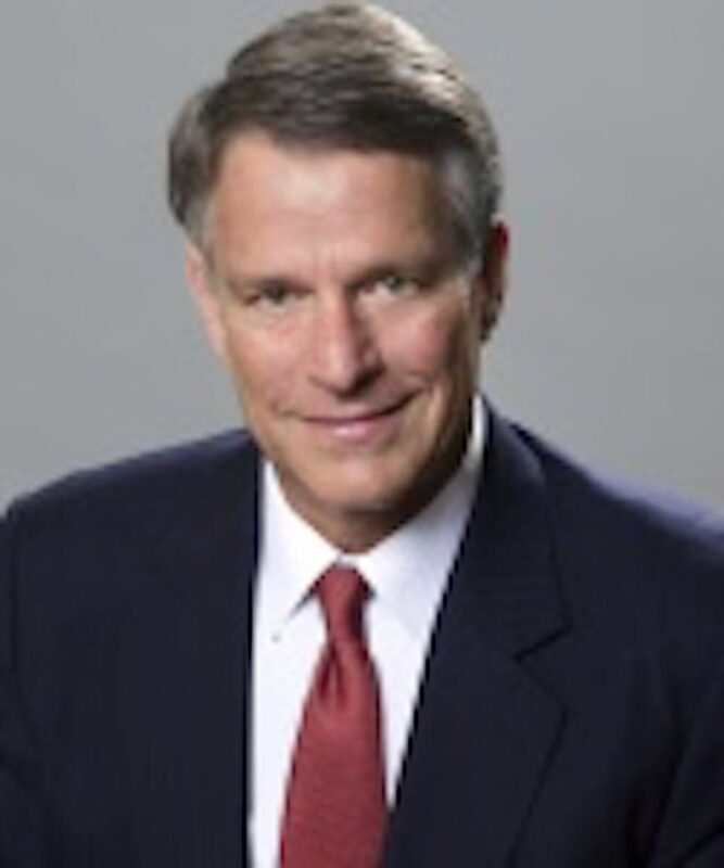 A man in a suit with a red tie, short gray hair, and a neutral expression, poses against a gray background.