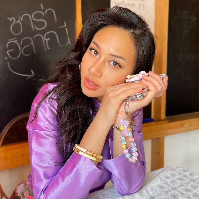 Person in a purple blouse holding a beaded accessory, sitting at a table with chalkboard background.