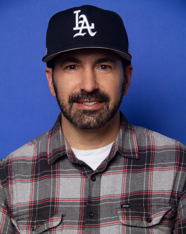 Man in a black LA baseball cap and plaid shirt smiles against a blue background.