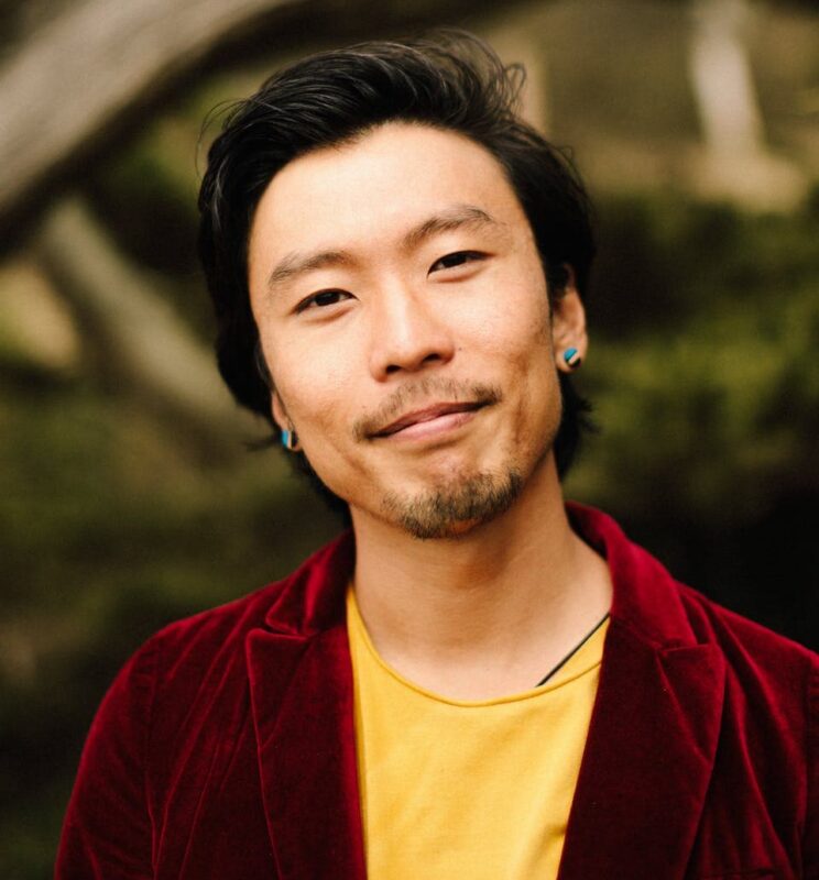 A person wearing a red jacket and yellow shirt smiles slightly, standing outdoors with blurred greenery and branches in the background.