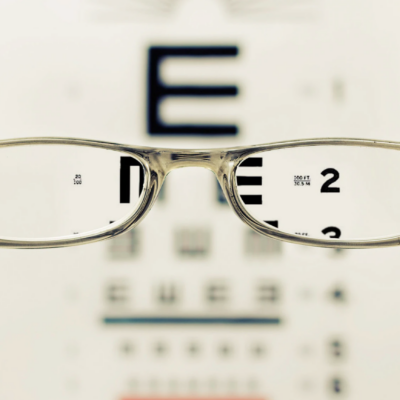 A person holds glasses focused on an eye chart, highlighting the "E" at the top, indicating improved vision with the lenses.