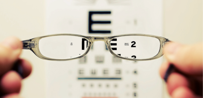 A person holds glasses focused on an eye chart, highlighting the "E" at the top, indicating improved vision with the lenses.