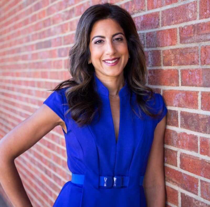 Person in a blue dress leaning against a red brick wall, smiling with hand on hip.