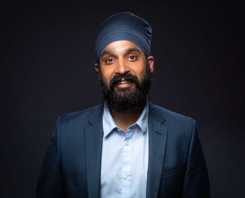 Man with a beard and turban, wearing a blue suit and light blue shirt, smiling against a dark background.