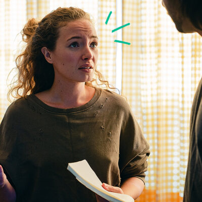 Woman holding a notepad looks concerned, talking to a man in a dimly lit room with yellow-checkered curtains.