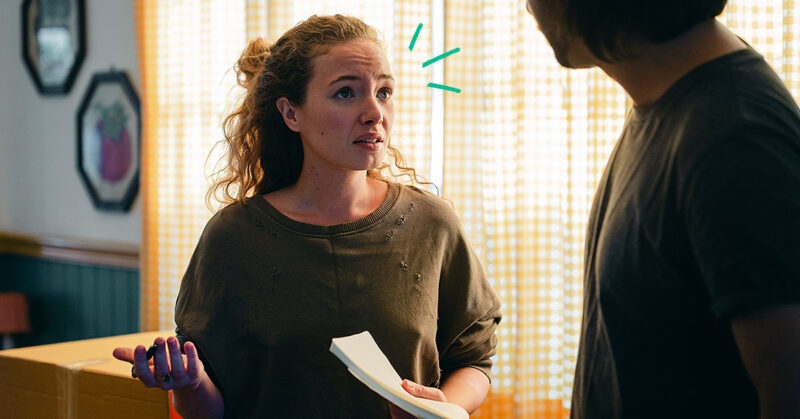 Woman holding a notepad looks concerned, talking to a man in a dimly lit room with yellow-checkered curtains.