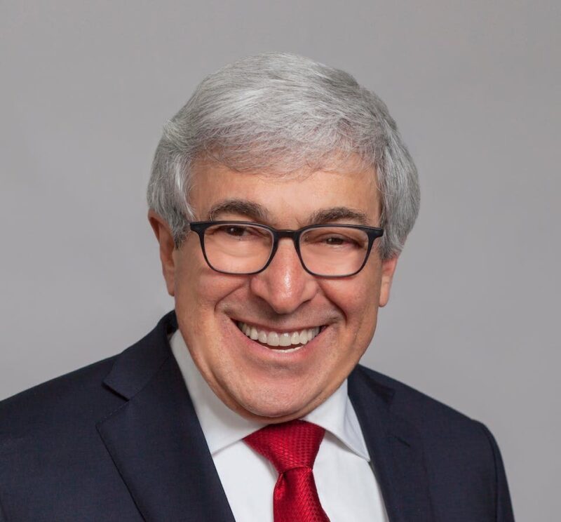 Smiling man with gray hair, glasses, a suit, white shirt, and red tie, against a plain background.