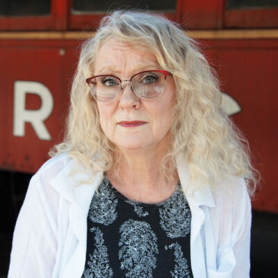 An older person with wavy blonde hair and glasses, wearing a patterned black shirt and white jacket, stands in front of a red background.