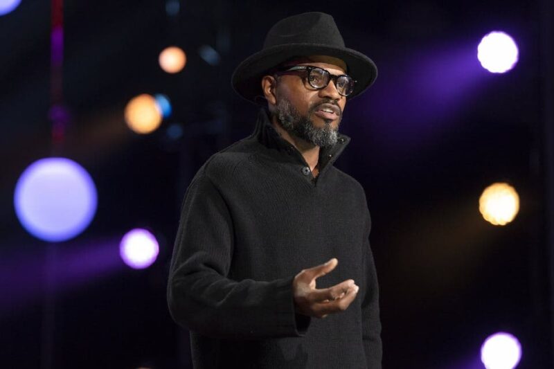 A man in a black hat and sweater speaks on a stage with colorful lights in the background.