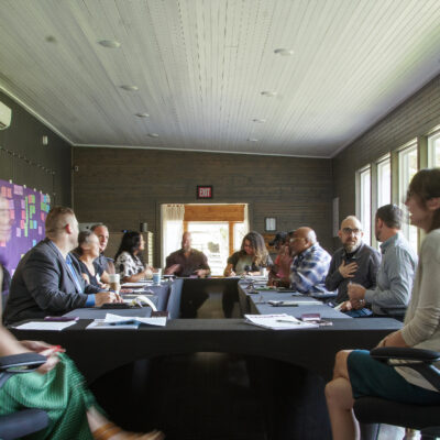 A group of people seated around a large rectangular table in a well-lit conference room, engaged in a meeting or discussion.