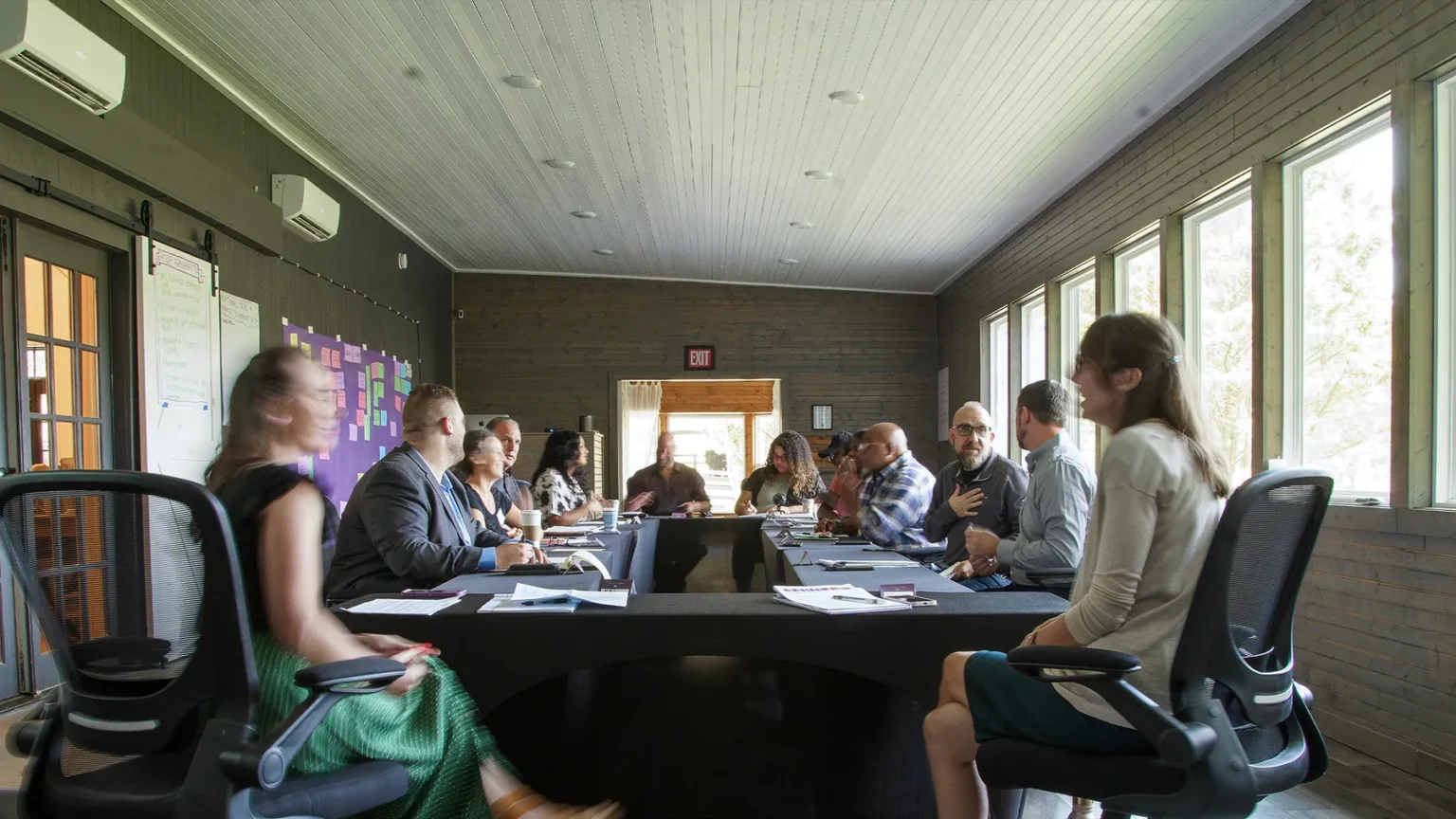 People sitting around a long conference table in a meeting room with large windows. Papers and laptops are on the table.