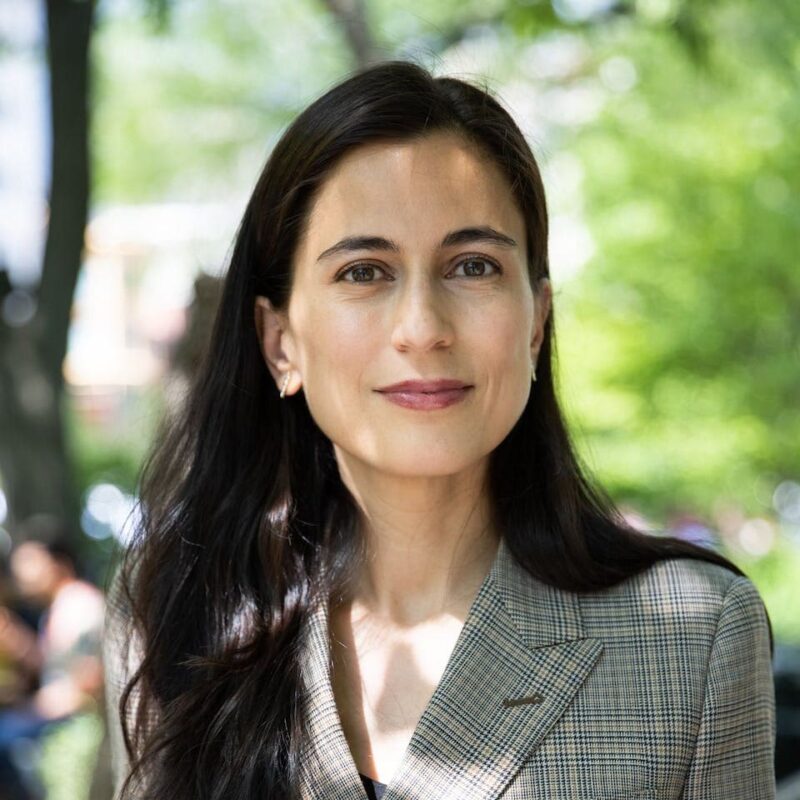 Woman with long dark hair wearing a plaid blazer, standing outdoors with trees and blurred people in the background.