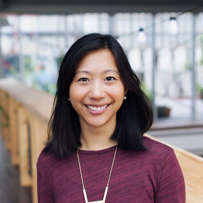 Smiling person with shoulder-length black hair and a maroon top, wearing a necklace, standing indoors. Blurred background with wooden elements.