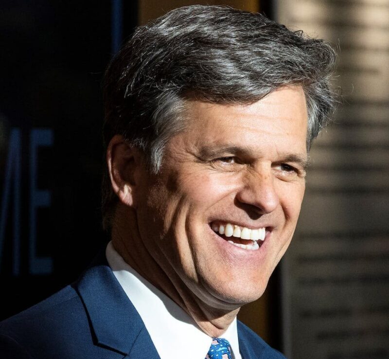 Smiling man in a suit with a blue patterned tie, standing indoors.