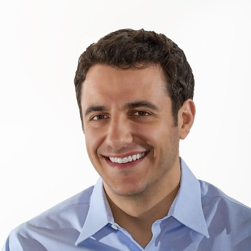 Smiling man with short dark hair wearing a light blue collared shirt against a plain white background.