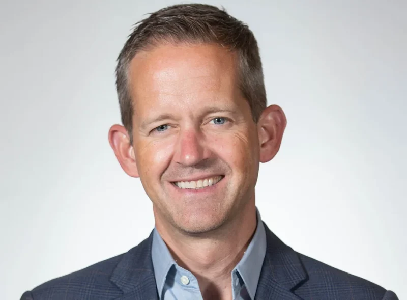 A smiling man wearing a dark blazer and light blue shirt, standing against a plain white background.