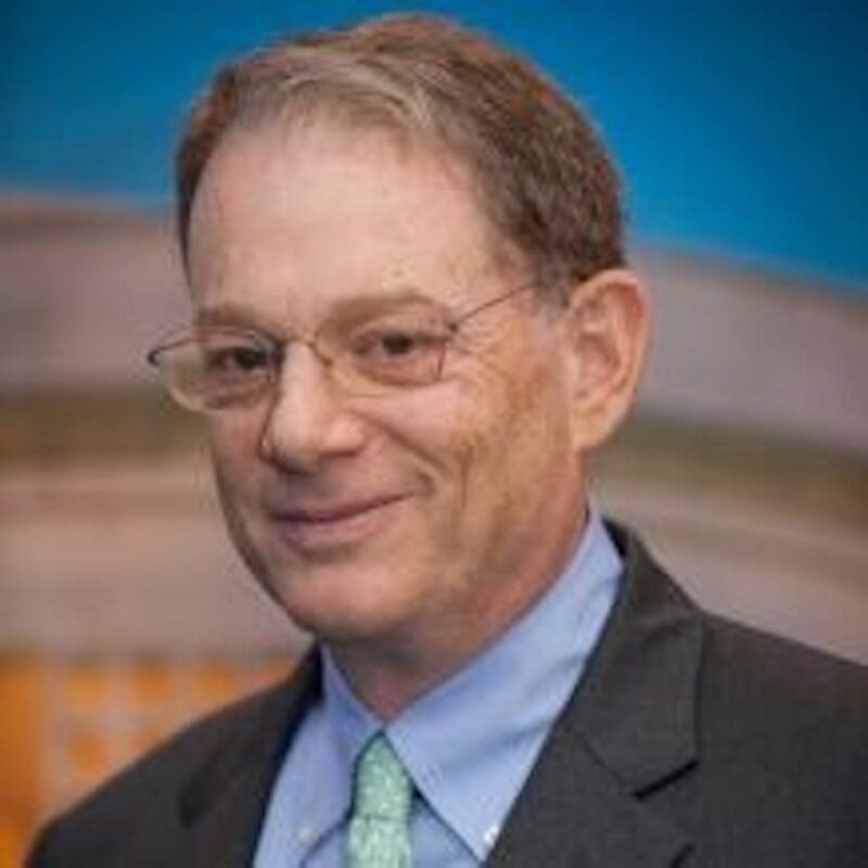 A man in glasses smiling, wearing a suit with a blue shirt and green tie, stands against a blurred background.