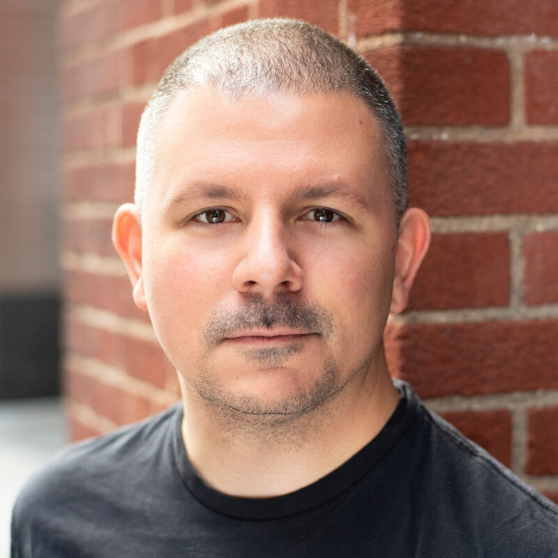 A person with a shaved head and short facial hair, wearing a black shirt, stands against a brick wall, looking directly at the camera.