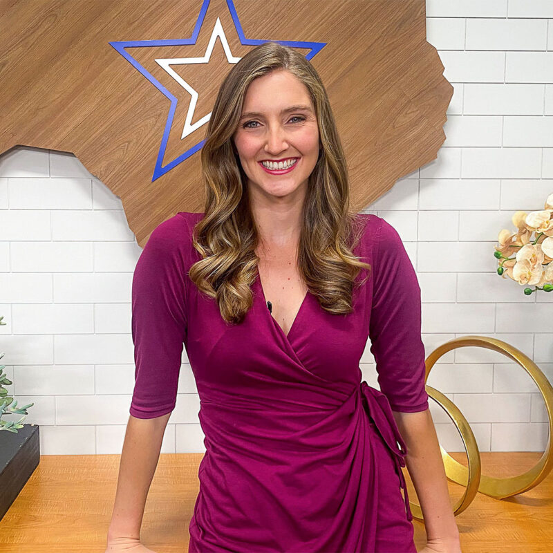 Woman in a maroon dress standing and smiling, with a wood star design on a white tile wall behind her.