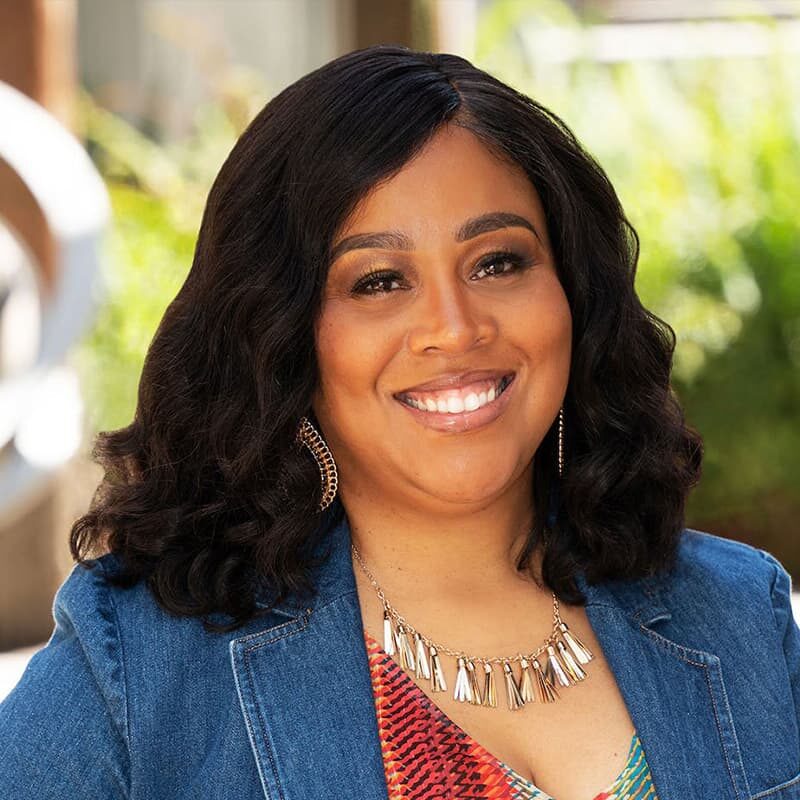 Smiling woman with long curly hair wearing a denim jacket and patterned top, standing outdoors with greenery in the background.