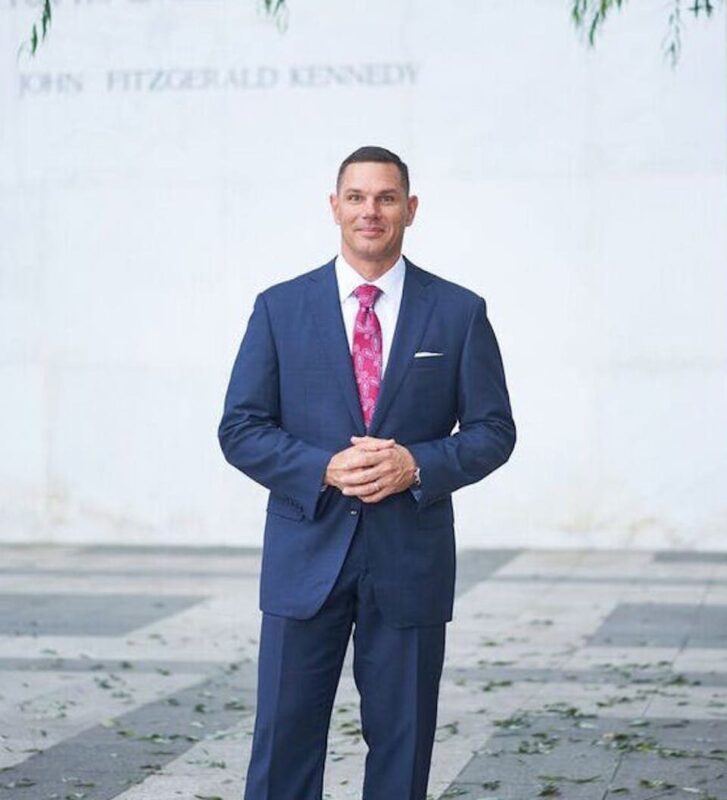 Man in a blue suit and pink tie stands outdoors on a paved area, with "John Fitzgerald Kennedy" visible on the wall behind him.