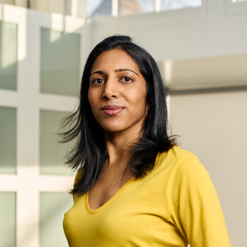 Person with long dark hair wearing a yellow top stands indoors.