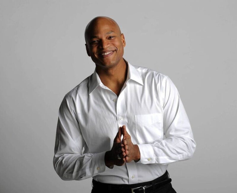 Smiling person in a white shirt clasping hands against a plain background.